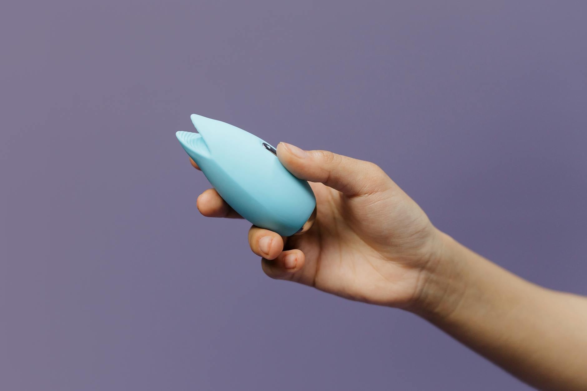 A blue silicone clitoral vibrator held in hand against a purple background, showing a tool for managing arousal mismatches in partnered sex.